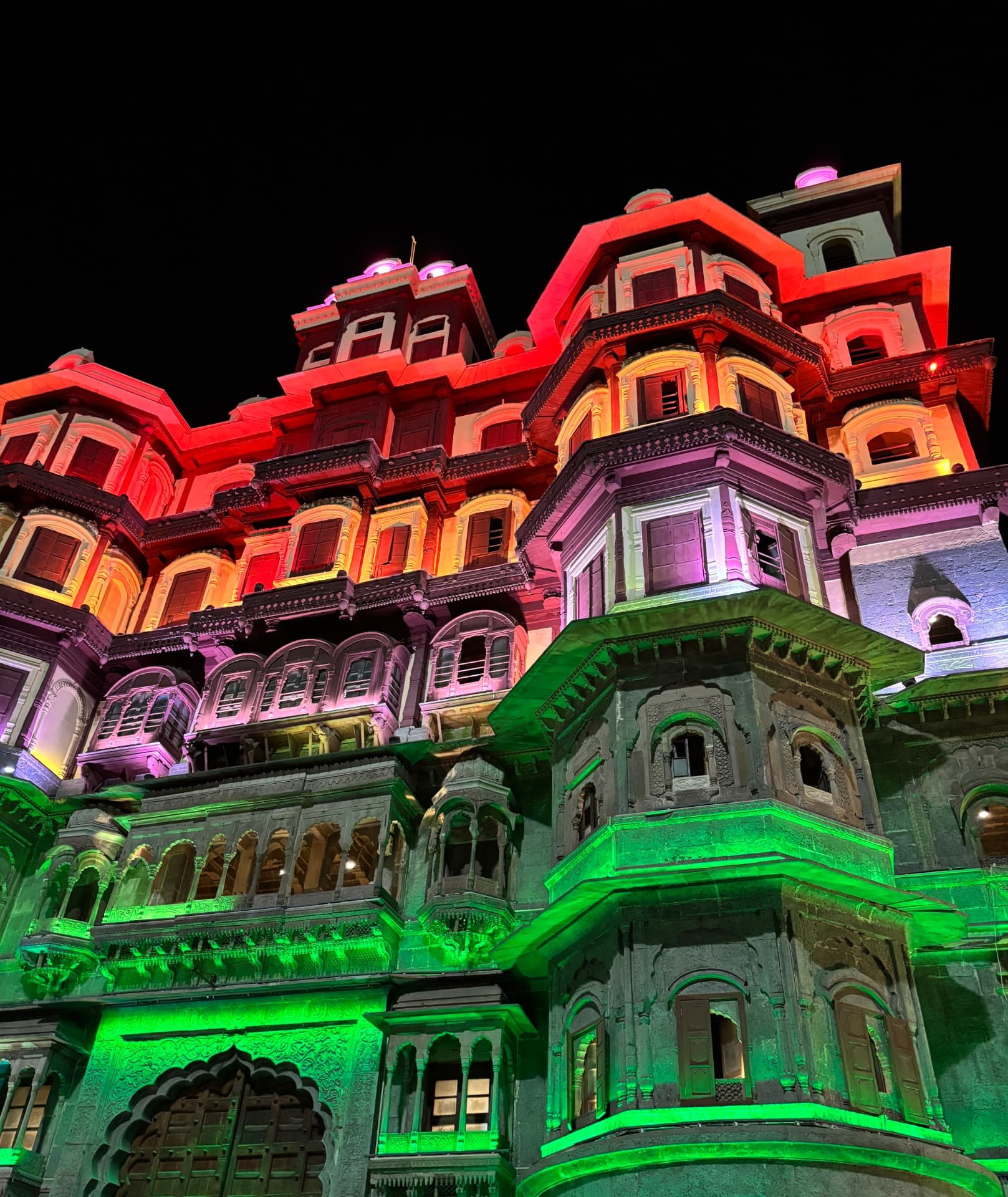 A historic Indian palace facade lit up in red, orange, pink and green lights against the night sky