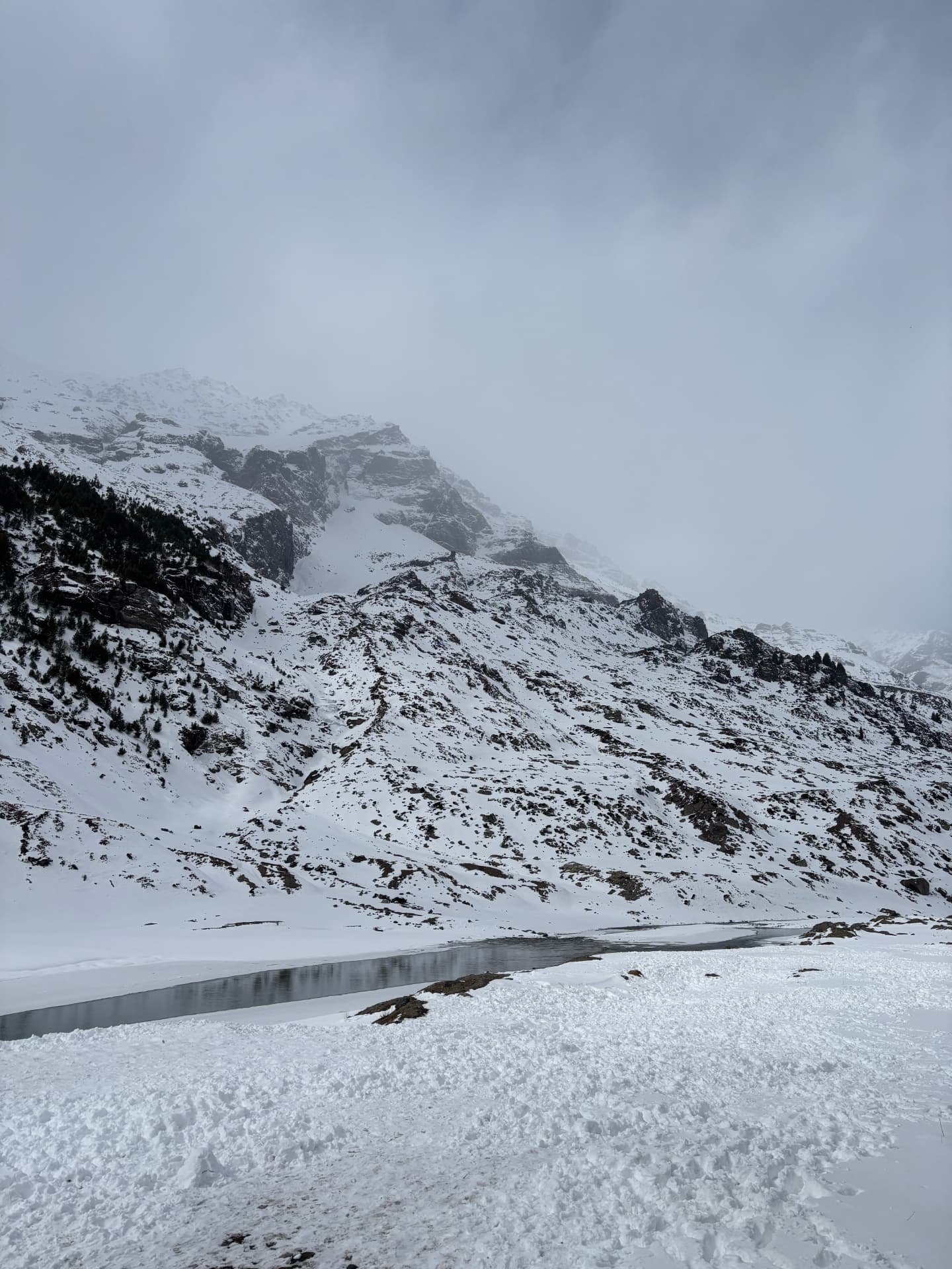 Snow-covered mountains beside a partially frozen stream under an overcast sky